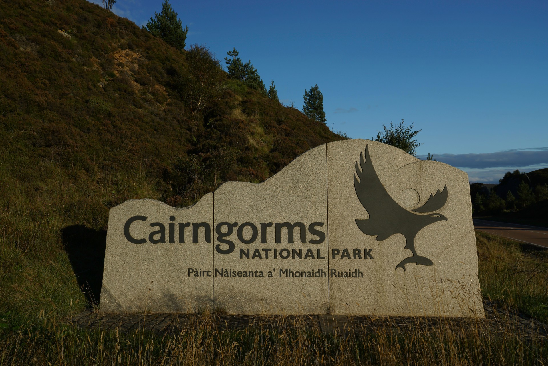 Cairngorms National Park Sign By Side Of A9 Road Lit By Golden Evening Sun Blue Sky And Hill With Greenery In Background No People 2GKXN8K